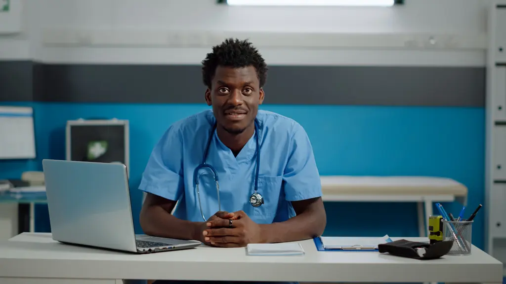 A nurse in blue scrubs smiling at his desk, with a laptop beside him